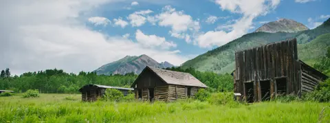 Old wooden structures stand in a green field with mountains in the background.