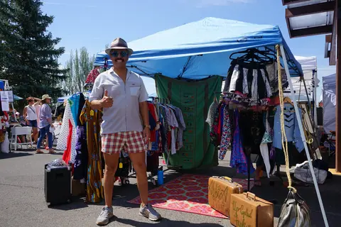 A man poses in front of a market tent selling clothing and vintage items.