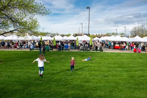 Children run on a grassy lawn near an outdoor market with many white tents and people.