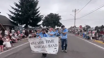 A marching band with a banner walks down a street in a parade.