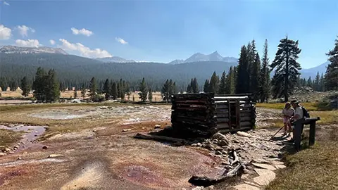 A log cabin stands in a mountain meadow with visitors nearby.