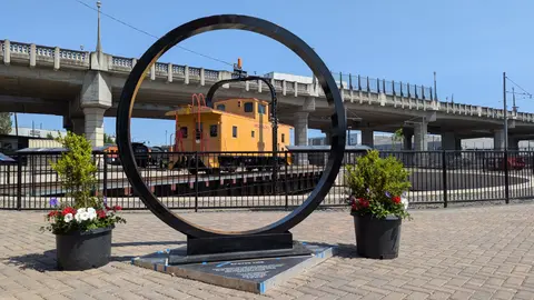 A large black ring sculpture stands on a brick-paved area with a yellow caboose in the background.