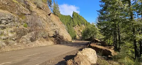 A dirt road winds through a mountainous landscape with rocky cliffs and pine trees.