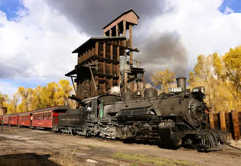 A steam train stands next to a wooden tower.