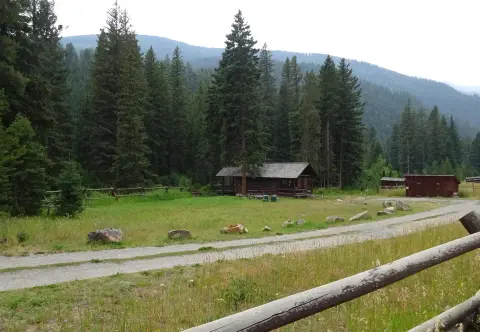 A log cabin stands in a clearing surrounded by a dense forest and mountains.