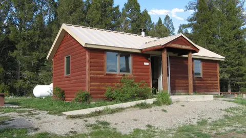 A red wooden cabin sits surrounded by trees and grassy ground.