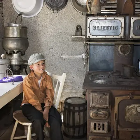 A young boy sits on a chair inside a rustic kitchen next to a vintage wood-burning stove.