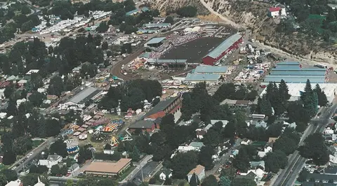 An aerial view shows a fairgrounds with a race track, grandstand, and exhibition buildings.