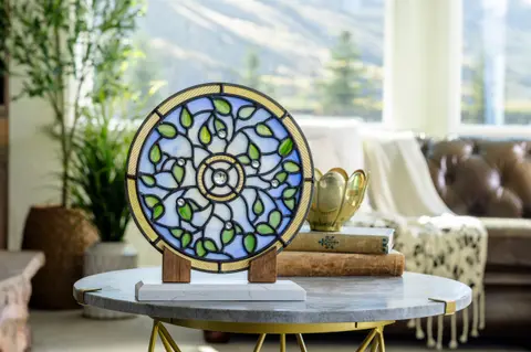 A round stained glass decor piece on a stand rests on a table indoors.