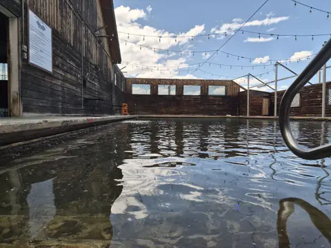 An outdoor swimming pool surrounded by wooden buildings and string lights under a cloudy sky.