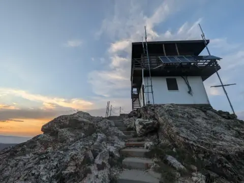 A fire lookout tower stands on top of a rocky peak at sunset.
