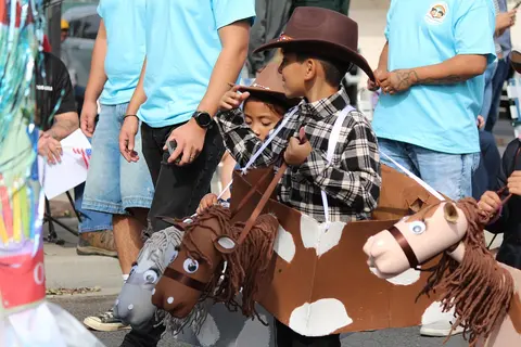 A child wearing a cowboy hat and plaid shirt sits inside a cardboard horse costume during a parade.