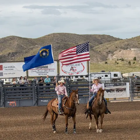 Two women ride horses while holding flags in a dirt arena.