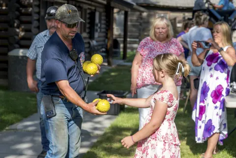 A man hands yellow balls to a girl outdoors while people watch.