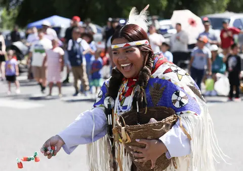 A smiling person in traditional regalia holds a basket and hands out candies at an outdoor gathering.