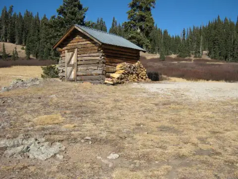 A small wooden cabin stands in a clearing with a pile of firewood stacked next to it.