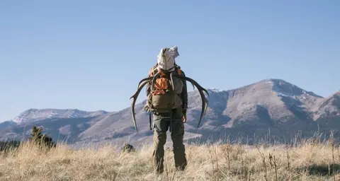 A hunter with antlers strapped to his backpack stands in a field with mountains in the background.