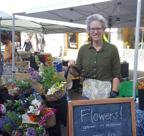 A woman smiles behind a flower stand at an outdoor market.