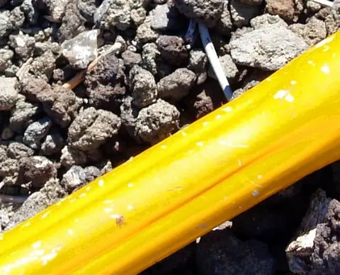 A close-up photograph of a yellow plastic pipe resting on gravel and rocks.