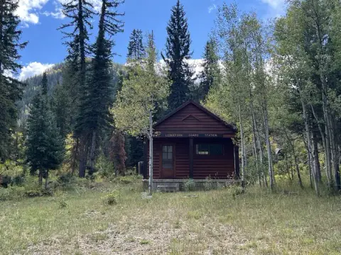 A log cabin sits nestled among tall trees beneath a blue sky.