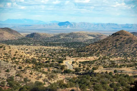 A scenic view of a winding road through dry, rolling hills and distant mountains under a cloudy sky.