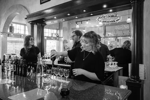 A black and white photograph showing people pouring wine at a bar interior.