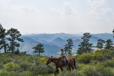 A person rides a horse on a hillside overlooking a mountain landscape.