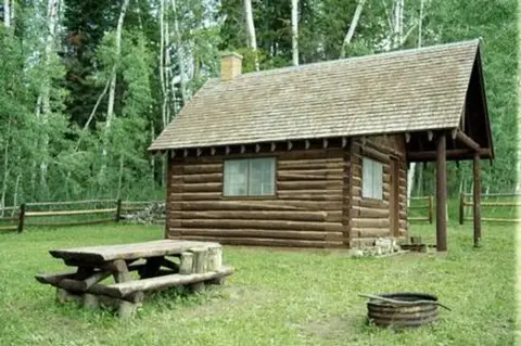 A log cabin stands in a grassy clearing surrounded by trees.