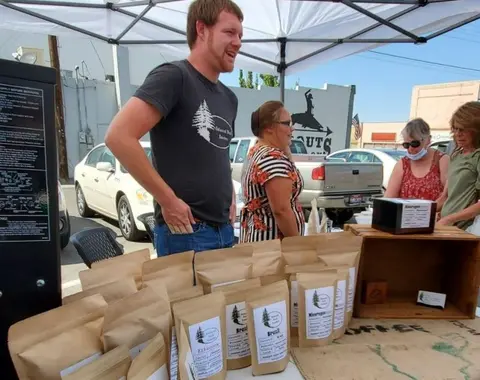 A man and a woman stand behind a table displaying packaged coffee beans at an outdoor market.