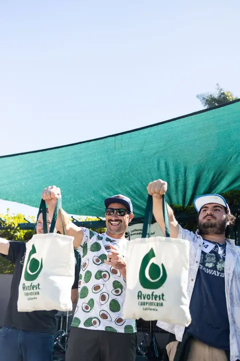 Three men hold up Avofest tote bags under a green canopy outdoors.
