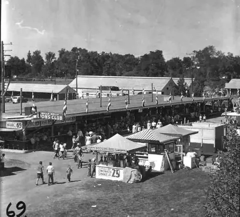 A historic black and white photo of a fairground with visitors and booths.