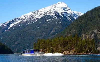 A boat cruises on a lake near a forested island with snow-capped mountains in the background.