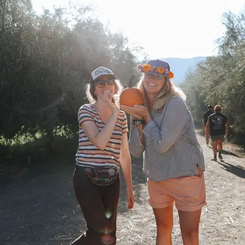 Two women smile on a dirt path with one holding a small pumpkin.