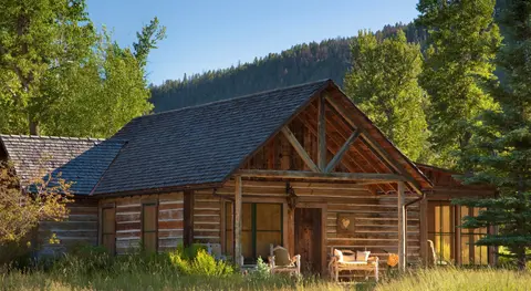 A rustic log cabin stands surrounded by trees with a mountain in the background.