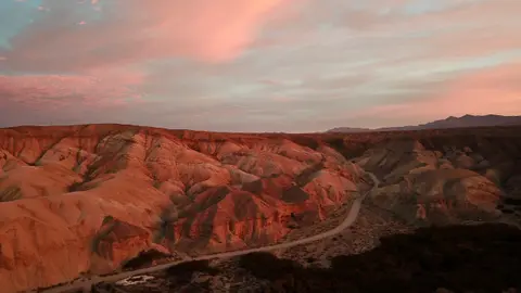 An aerial view of a winding desert road through barren hills under a colorful sunset sky.