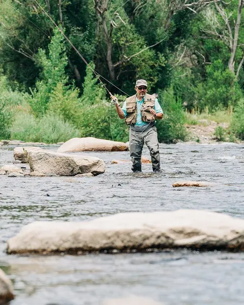 A man fly fishes in a river surrounded by trees and rocks.
