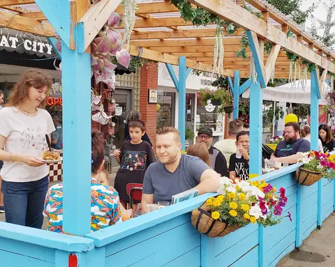 People dining in an outdoor seating area with blue wooden railings and a wooden trellis.