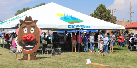 A large white tent with a potato mascot cutout and people gathered underneath on a grassy field.