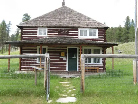A rustic two-story log building with a covered porch stands in a grassy clearing.