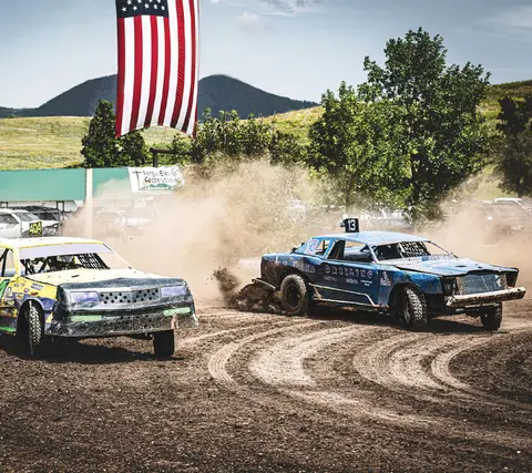 Two stock cars race on a dirt track kicking up dust beneath a large American flag.