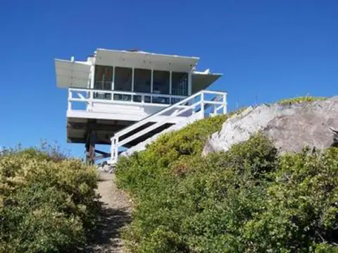 A white lookout tower stands on a hill surrounded by green bushes under a clear blue sky.