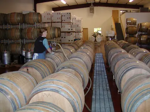 A woman works in a large warehouse filled with stacked wooden barrels.