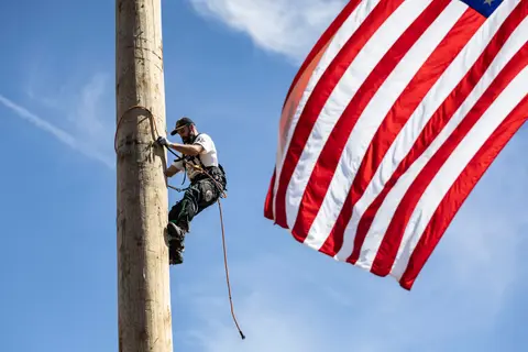 A lineman works on a wooden pole with an American flag nearby.