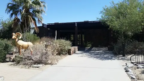 A walkway leads to the entrance of a building surrounded by desert landscaping and a bighorn sheep sculpture.