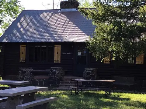 A log cabin with a corrugated metal roof stands among trees with picnic tables in the foreground.