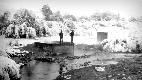 Historic black and white photograph showing two people standing on a concrete structure near a steaming stream in a snowy landscape.