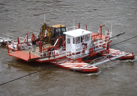 An aerial view shows a red and white ferry boat carrying a backhoe across a river.