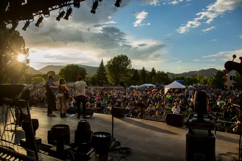 Musicians perform on an outdoor stage before a large audience seated on the grass.