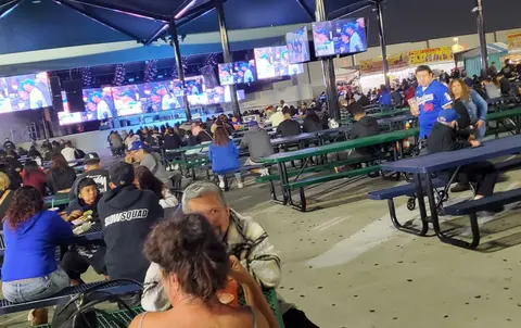 People sit at outdoor picnic tables under a pavilion watching sports on multiple screens.