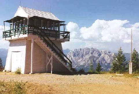 A fire lookout tower stands on a mountain peak with distant rugged mountains and a blue sky in the background.